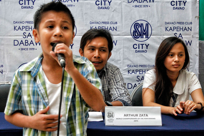 SHOWCASING WORLD-CLASS TALENT.  Lloyd Montebon, 11, of Angliongto village in Davao City belts out a song during Monday’s SM Kapehan.  He’s one of the 30 Mindanaoans who are hoping to bag a gold medal from the World Championships of Performing Arts competition in Hollywood, USA.  Behind him are Director Arthur Zata (left) and fellow qualifier Jizelle Dea Formilleza.  (davaotoday.com photo by Medel V. Hernani)