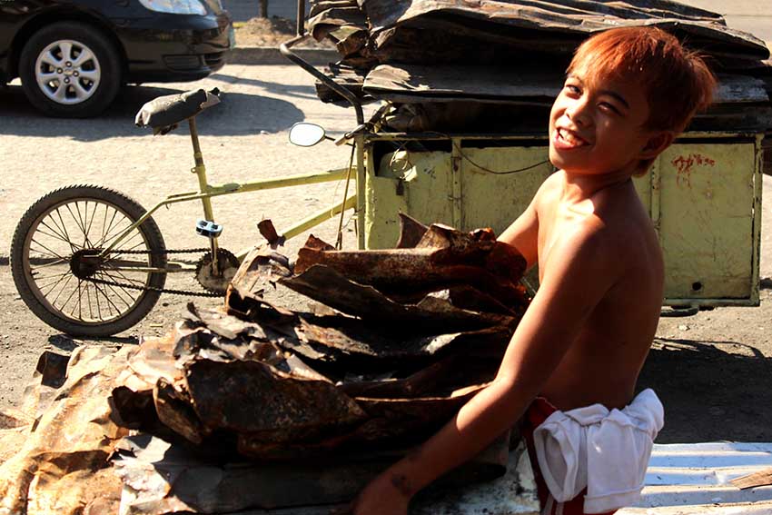 A boy flashes his smile to the lens of the camera while carrying scrap galvanize iron roof.